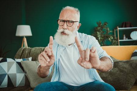 Senior man with white beard sits on a cozy living room couch in a warm home interior expressing friendly gestures creating a light hearted family moment at homeの写真素材