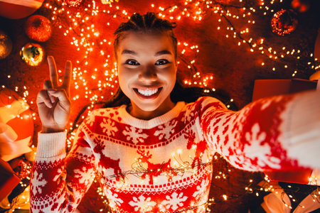 Playful holiday moment as a joyful woman in a red white sweater takes a selfie surrounded by warm lights gifts and a festive treeの写真素材