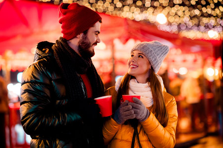 Cheerful winter couple at festive outdoor market sharing warm drinks under twinkling lightsの写真素材