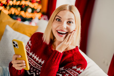 Funny Christmas moment as happy blonde woman in red sweater chats on her phone at home with festive lights and tree aroundの写真素材