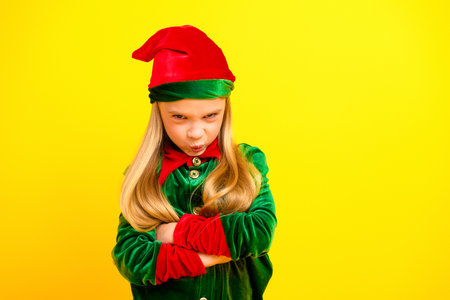 Festive girl dressed as a stylish elf in green and red costume on yellow backdrop, expressing...