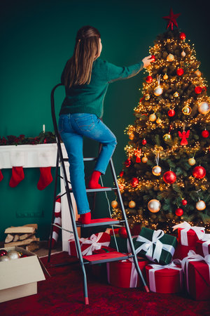 A cheerful helper climbs a ladder to decorate a tall Christmas tree in a cozy living room full of lights gifts and warm holiday atmosphere perfect for a family celebrationの写真素材