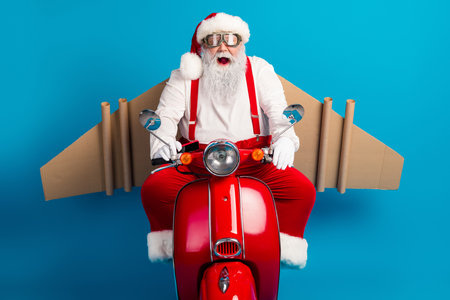 Santa on a bright red scooter with cardboard wings smiles for a festive photo shoot against a blue studio backdrop during magical Christmas seasonの写真素材