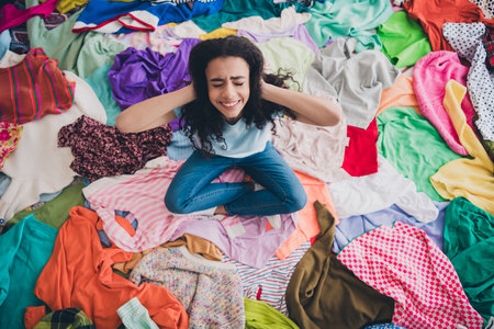 High angle view full size photo of young girl hands touch head sit pile stack clutter clothes apartment indoorsの写真素材