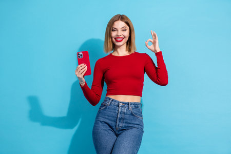 Beautiful young woman in casual outfit holding phone against a blue background, smiling joyfully...