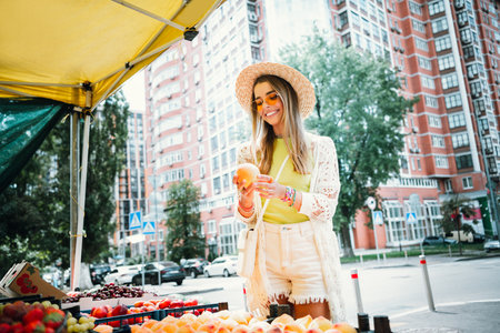 A fashionable young woman at a sunny city market wearing a straw hat smiles while picking fruit under a yellow canopyの写真素材