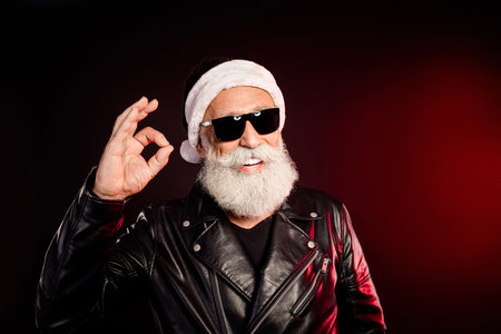 Santa rocker in leather jacket with white beard wears black sunglass and gives the ok gesture in a festive portraitの写真素材