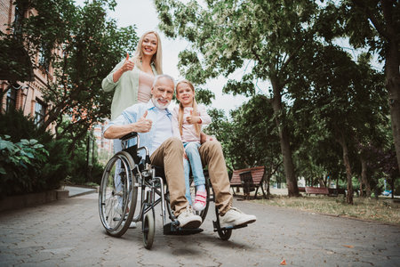 Happy family day out in the park joyful grandpa in wheelchair with daughter and granddaughter smiling and giving thumbs up for care and recoveryの写真素材