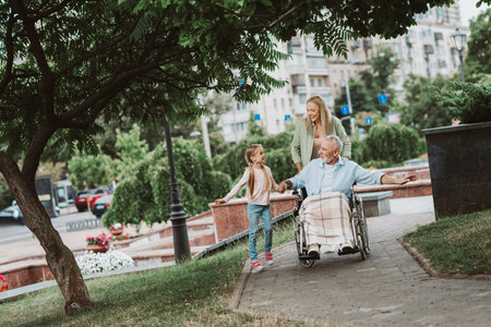 Funny heartwarming park day with grandpa in wheelchair smiling with daughter and granddaughter as loving caregiver joinsの写真素材