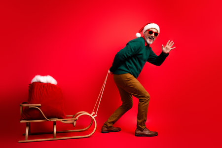 Funny santa pulls a sleigh with a red sack as a cheerful man in a green sweater waves in a bold red studioの写真素材