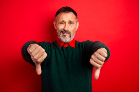 Funny older man in green sweater poses with thumbs down against bright red background as a playful holiday stock photo conveying disapproval mood and warm festive vibes for christmas seasonの写真素材