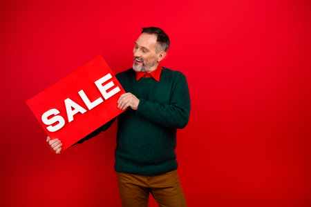 Funny man with sale sign wearing green sweater and bow tie stands against bright red background promoting holiday cheerの写真素材