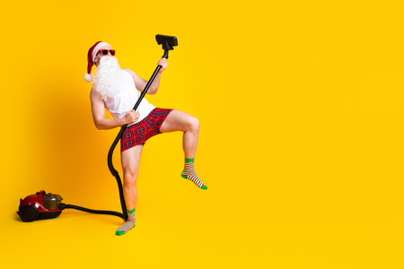 Santa in funny pajama poses with vacuum on bright yellow background for holiday fun and festive chaos new year ready family vibeの写真素材