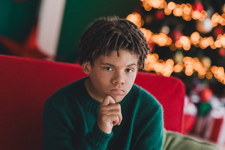 Thoughtful teenage boy sitting indoors near a beautifully decorated Christmas tree during the festive season.の写真素材