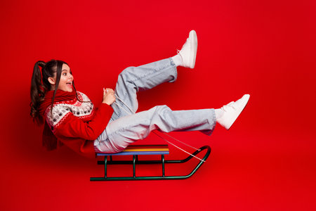 Joyful young woman on a red sled wearing sweater and scarf against bright red background enjoying Christmas season fun and festive holiday vibes in a playful winter momentの写真素材