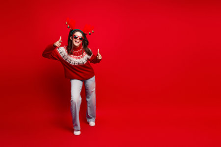 Happy teen girl in red christmas sweater with reindeer headband posing against bold red background celebrating festive season and holiday joy with thumbs up and playful fashionの写真素材