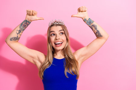 Young woman with crown in blue top poses against pink background with arms raised and thumbs down smilingの写真素材