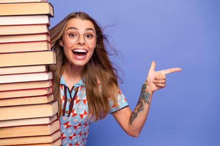 Young woman with stack of book smiling and pointing against purple backgroundの写真素材