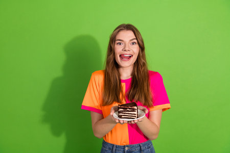 Young woman with brown hair smiling holds chocolate cake slice in colorful neon shirt against bright...