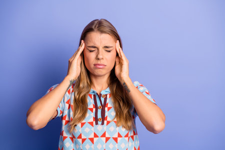 Young woman with headache holding temples against a purple background wearing a patterned casual...
