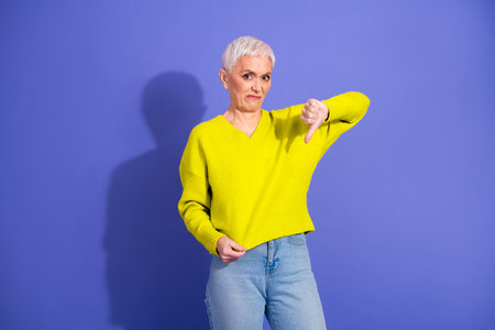 Senior woman in bright yellow sweater gives thumbs down in a bold fashion and lifestyle photo against blue backgroundの写真素材