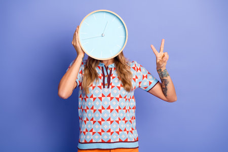 Young woman covers her face with a clock while flashing a peace sign against a vibrant studio...