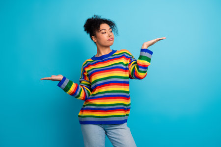 Young woman in a colorful striped sweater stands against a blue background with open hands