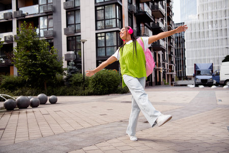 Young traveler woman with pink backpack and lime green vest enjoys sunny city walk with headphonesの写真素材