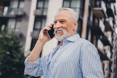 Senior man on a city street talking on his phone smiling in a striped shirt with modern apartment...