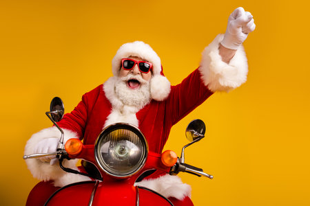 Santa on a red motorcycle rides against a bright yellow backdrop with sunglass and a triumphant raised fist for holiday cheerの写真素材