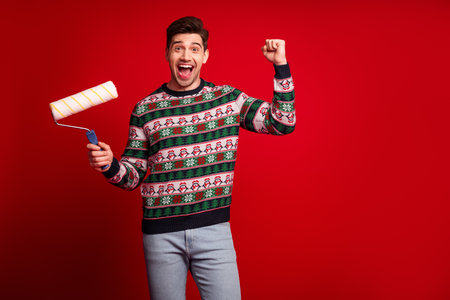 Joyful man in a Christmas sweater celebrates the season as he paints a bold red background with a roller and looks ready for holiday funの写真素材