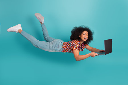 Young mixed race woman with curly hair wears a patterned shirt and jeans flies through the air holding a laptop against a blue background smiling and joyfulの写真素材