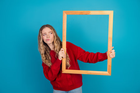 Charming young woman in a red sweater holding a wooden frame against a bright blue background showcasing casual styleの写真素材