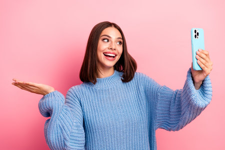 Young woman in blue knit sweater smiles while taking a selfie with a blue phone against a pink backgroundの写真素材