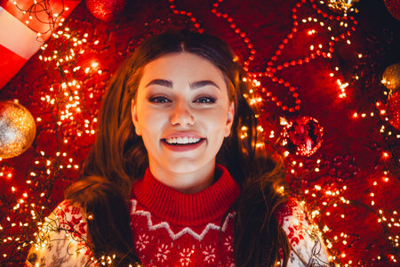 Cheerful woman in red christmas sweater surrounded by warm lights and festive decorations at home during xmas celebrationの写真素材