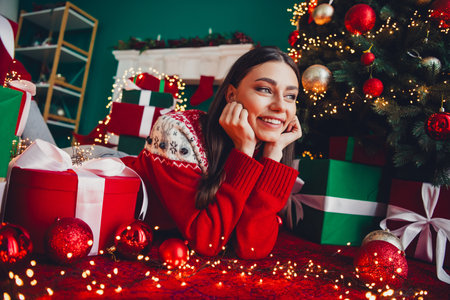 Cozy Christmas joy as a young woman in a red sweater dreams beside a lit tree and wrapped giftsの写真素材