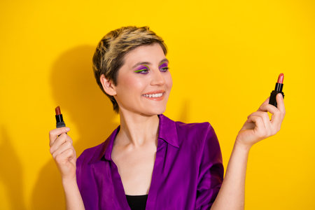 Young woman in purple blouse smiles while holding lipstick tubes against a bright yellow background showcasing glamour fashion and beautyの写真素材