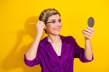 Young woman in a vivid purple blouse applies makeup and checks her reflection in a round mirror against a bold yellow backgroundの写真素材