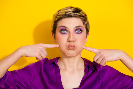 Playful young woman with short blonde hair in a purple blouse makes a funny pout while pointing at her cheeks against a bright yellow backgroundの写真素材