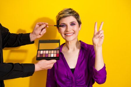 Young woman with short blonde hair smiles as makeup artist applies eye shadow and shows peace sign against a bright yellow backgroundの写真素材