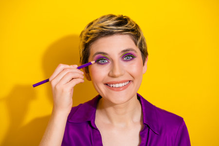 Young woman with bold purple eye makeup smiles as she applies liner against a vibrant yellow backgroundの写真素材