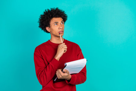 Thoughtful young man in red sweater holds notebook and pencil while looking upward in a bright teal background exploring ideas and study conceptsの写真素材