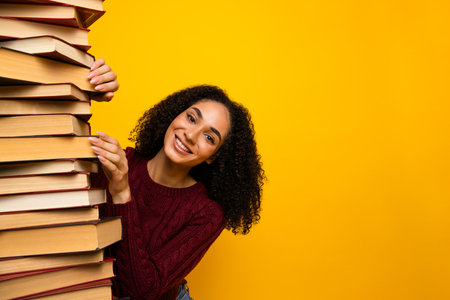 Young mixed race woman smiles while stacking a tall pile of book against a bright yellow background for study and discoveryの写真素材