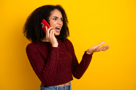 Young mixed race woman on yellow background talking on smartphone and gesturing with open hand in a casual shopping lifestyle advertising sceneの写真素材