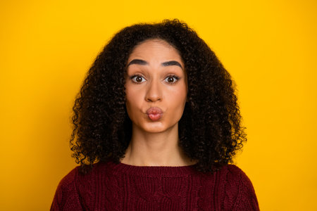 Young mixed race woman with curly hair in a burgundy sweater making a playful kissy face against a bright yellow background for stockの写真素材