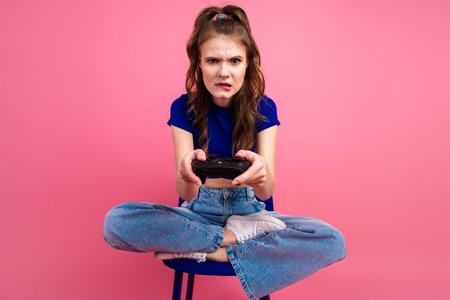 Young woman with a game controller sitting cross-legged against pink background, expressing...