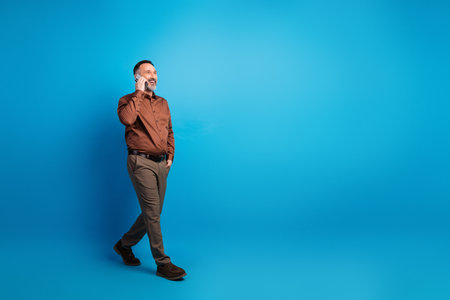 Professional mature businessman on blue studio background talking on phone while walking confidently in smart casual shirt and tailored pantsの写真素材