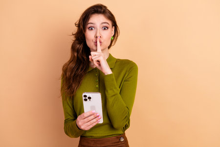 Young woman with finger to lips holding smartphone in a casual stylish pose against a beige backgroundの写真素材