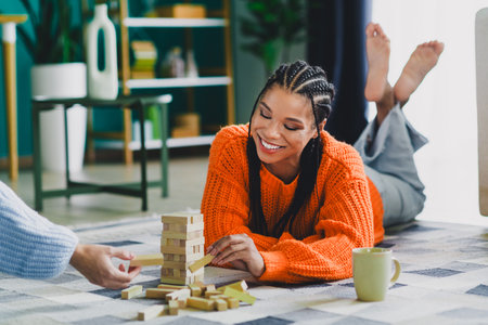 Young woman in a bright orange knit sweater enjoys a cozy home moment as she builds a wooden block tower with a friend on a sunny living room floor todayの写真素材