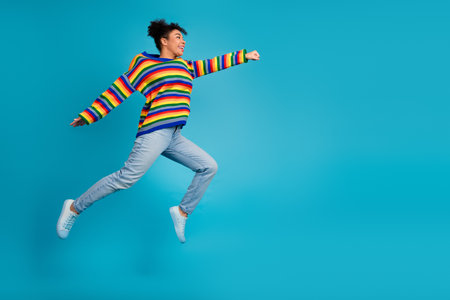 Young woman leaps in a colorful striped sweater against a blue background capturing joy style and carefree energyの写真素材